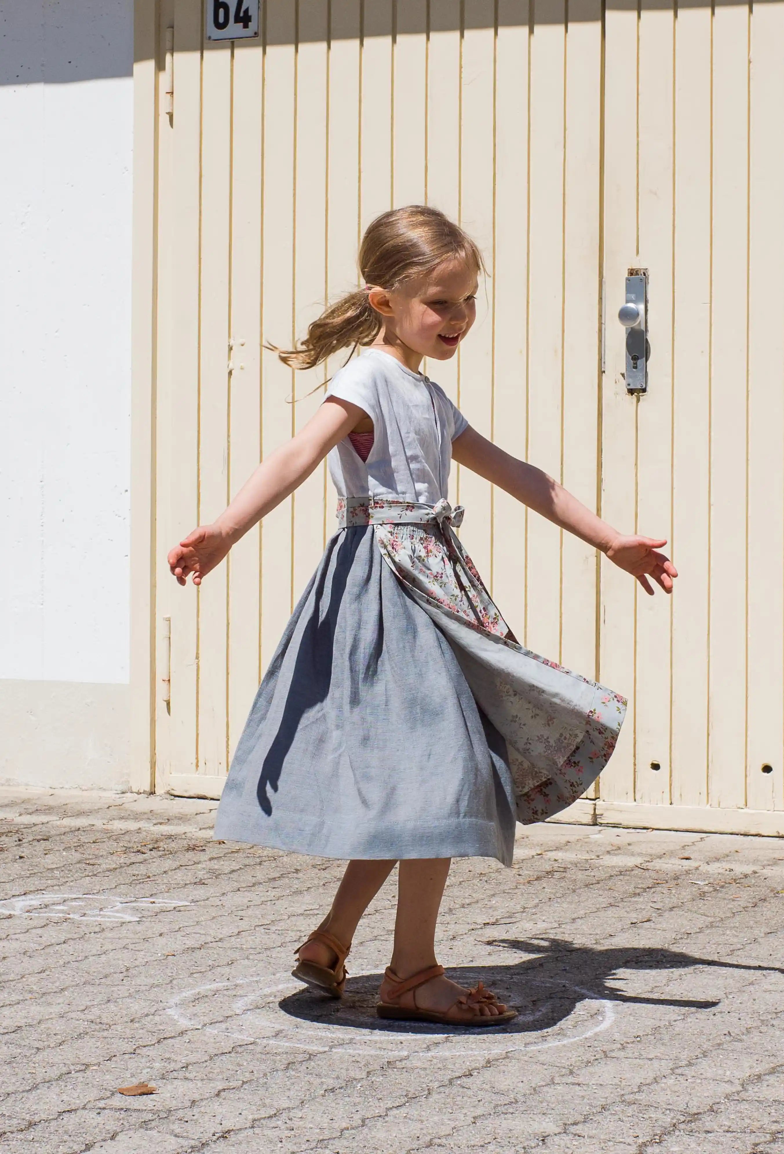 Zirbenfüxlein Kinderdirndl Saphir aus Leinen mit hellblauer Schürze mit Blumenmuster, schwungvolle Drehung mit ausgestreckten Armen in Frontansicht vor urbanen Garagentoren, lebendig umgesetzt von Zirbenfüxlein.