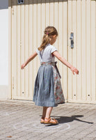Zirbenfüxlein Kinderdirndl Saphir aus Leinen mit hellblauer Schürze mit Blumenmuster, Kind in Drehbewegung in Rückansicht vor urbanen Garagentoren, lebendig umgesetzt von Zirbenfüxlein.