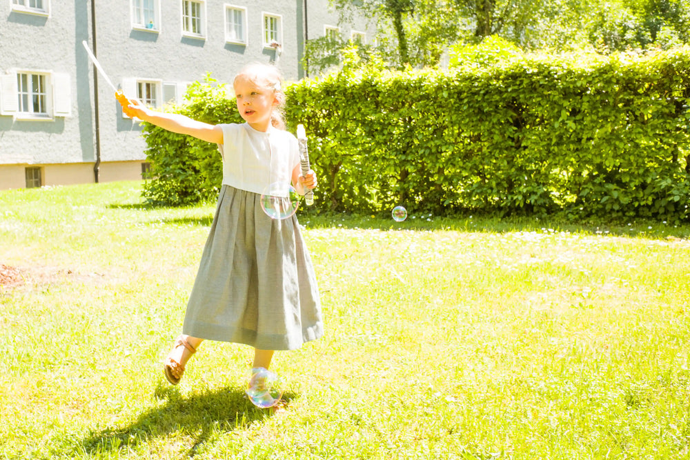 Kinderdirndl in Saphirblau – Frontansicht in Bewegung auf einer Hinterhofwiese, umgeben von Seifenblasen, lebendig inszeniert von Zirbenfüxlein