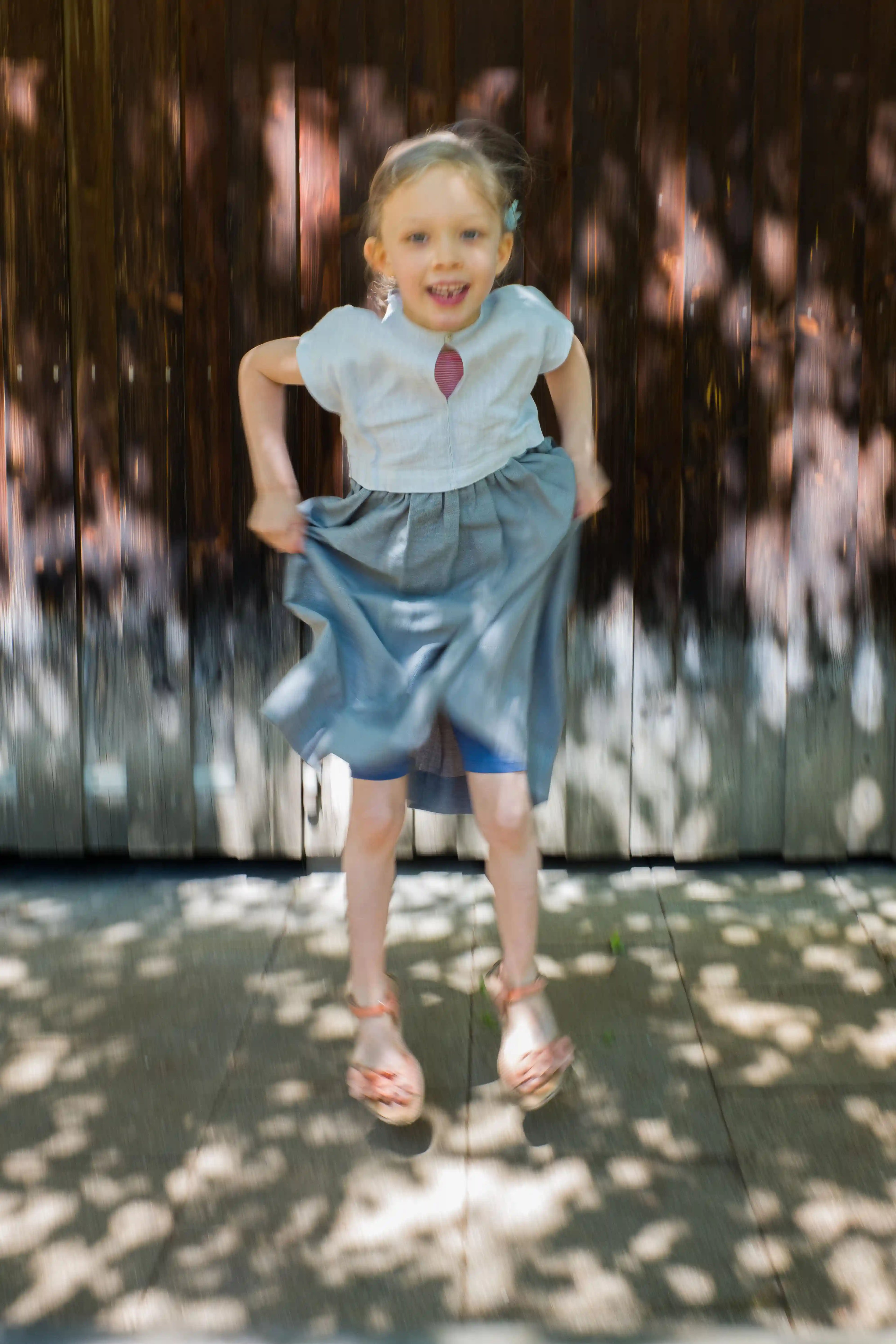 Kinderdirndl in Saphirblau – nach oben springend vor einer Altholzwand im urbanen Setting, markantes Licht- und Schattenspiel, dynamisch inszeniert von Zirbenfüxlein