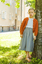 Zirbenfüxlein Kinderdirndl Saphir mit terrakottafarbener Strickjacke, Kind in Frontansicht auf einer Wiese an einen Baum angelehnt in urbaner Hinterhofszene, klar gestaltet von Zirbenfüxlein.