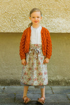 Zirbenfüxlein Kinderdirndl Saphir mit terrakottafarbener Strickjacke und hellblauer Schürze mit Blumenmuster, Frontansicht vor neutraler urbaner Fassade, klar interpretiert von Zirbenfüxlein.