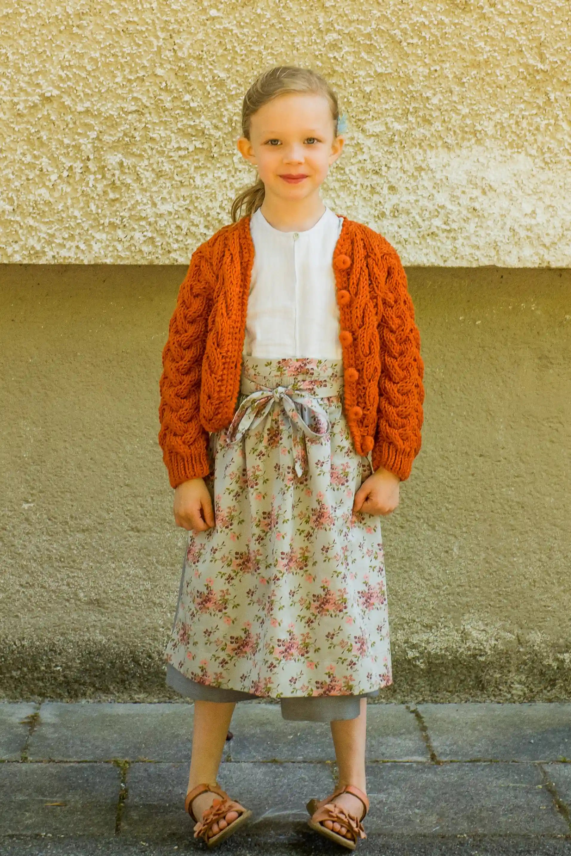 Zirbenfüxlein Kinderdirndl Saphir mit terrakottafarbener Strickjacke und hellblauer Schürze mit Blumenmuster, Frontansicht vor neutraler urbaner Fassade, klar interpretiert von Zirbenfüxlein.