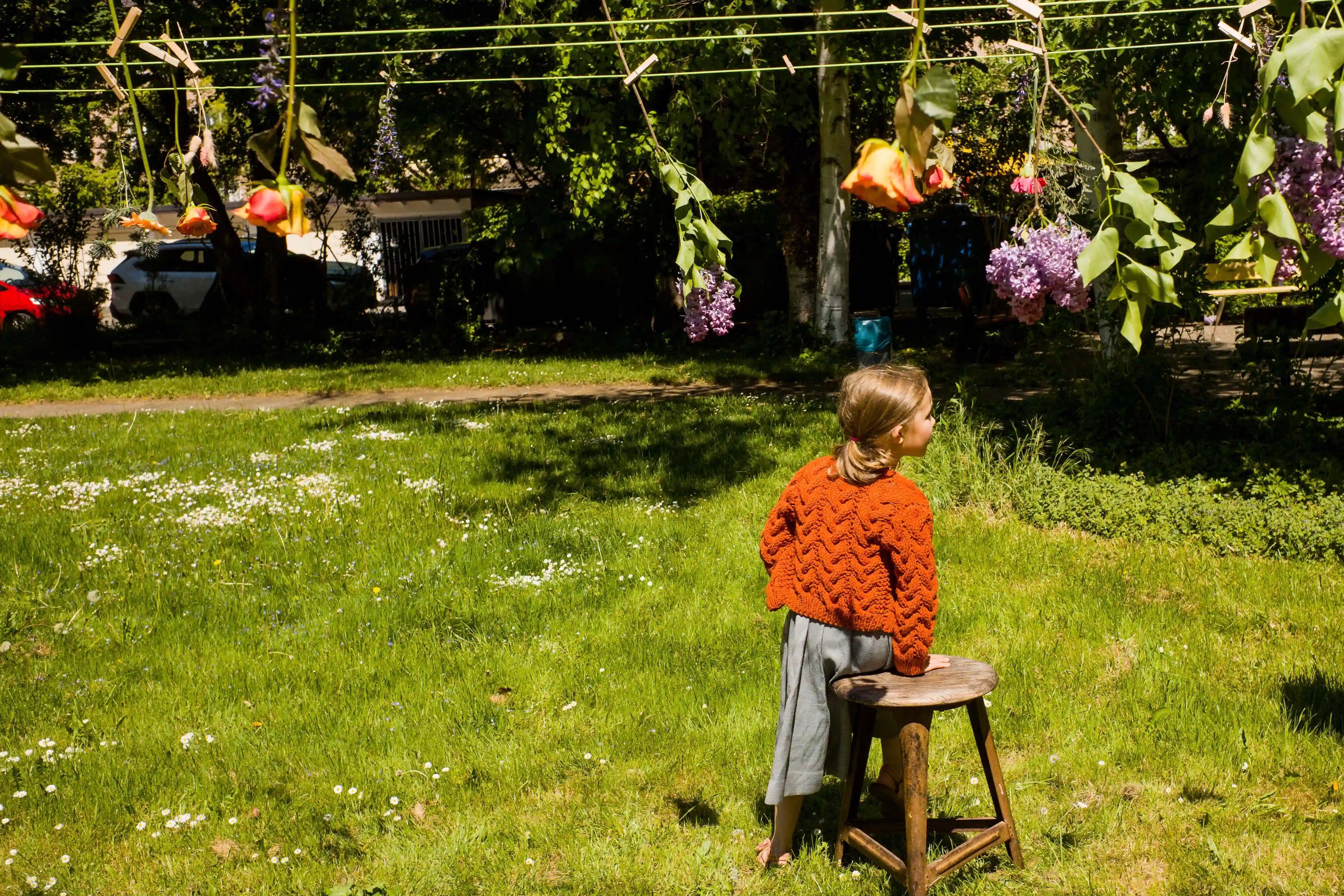 Zirbenfüxlein Kinderdirndl Saphir mit terrakottafarbener Strickjacke, Rückansicht eines sitzenden Kindes auf altem Hocker auf einer Hinterhofwiese, darüber bunte Blumen an einer Wäscheleine, atmosphärisch inszeniert von Zirbenfüxlein.