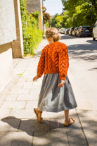 Zirbenfüxlein Kinderdirndl Saphir mit terrakottafarbener Zopf-Strickjacke, Rückansicht eines Kindes in Drehbewegung mit Blick nach unten auf dem Gehsteig in sonniger urbaner Straßenszene, modern interpretiert von Zirbenfüxlein.
