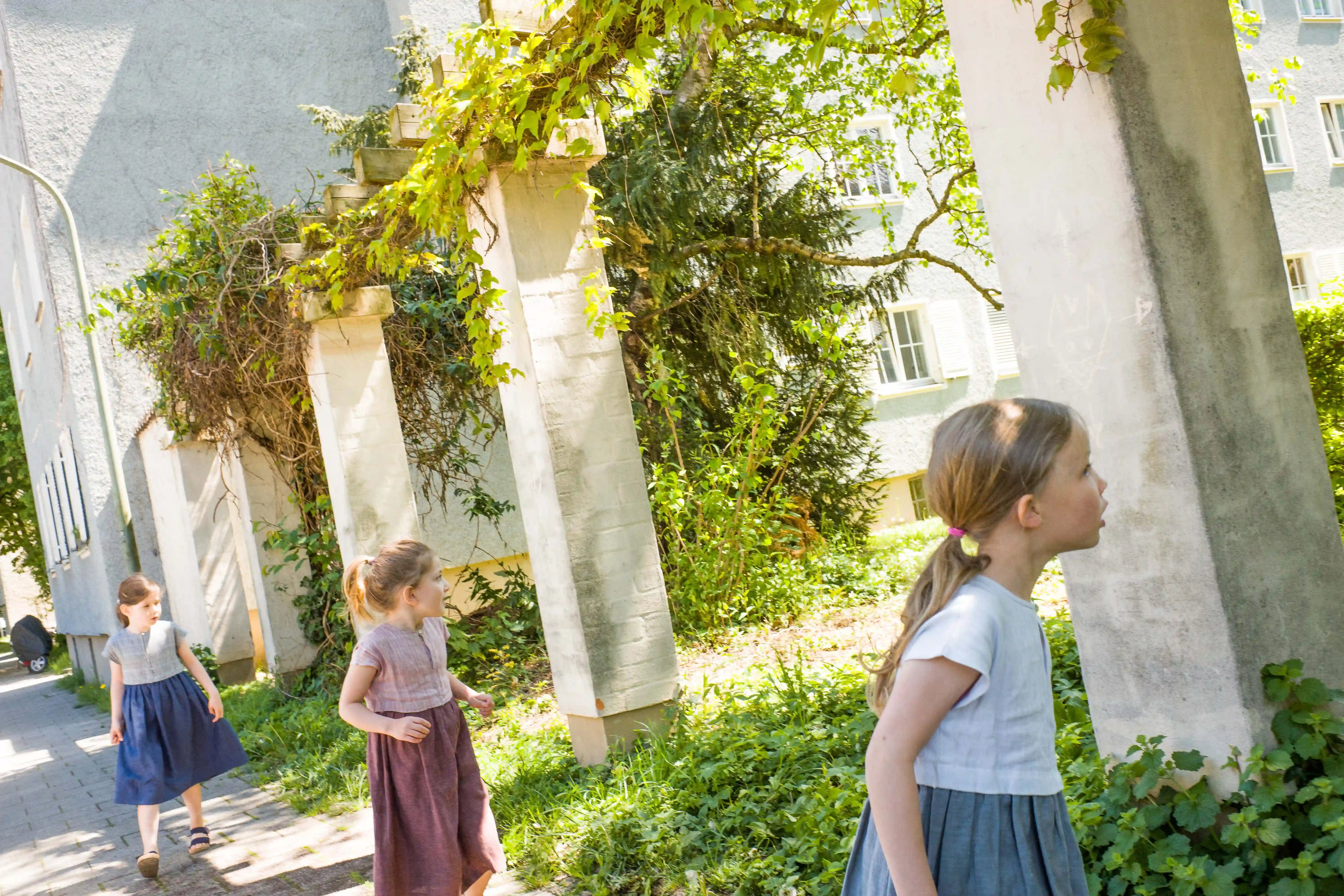 Zirbenfüxlein Kinderdirndl in Saphir, Tintenblau und pinker Beere, drei Kinder suchend in grüner Hinterhofszene dargestellt, zurückhaltend umgesetzt von Zirbenfüxlein.