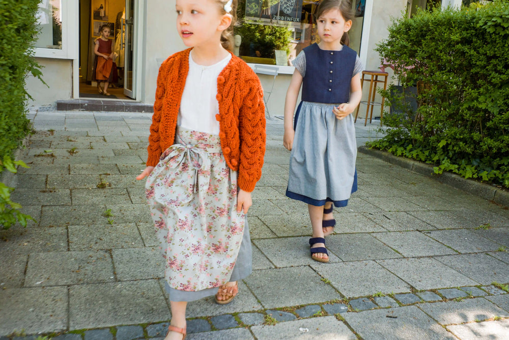 Zirbenfüxlein Kinderdirndl in Saphir und Tintenblau mit Strickjacke, drei Kinder in Bewegung beim Verlassen des Zirbenfux Ladens, zwei bereits draußen, eines tritt gerade durch die Tür, offen dokumentiert von Zirbenfüxlein.