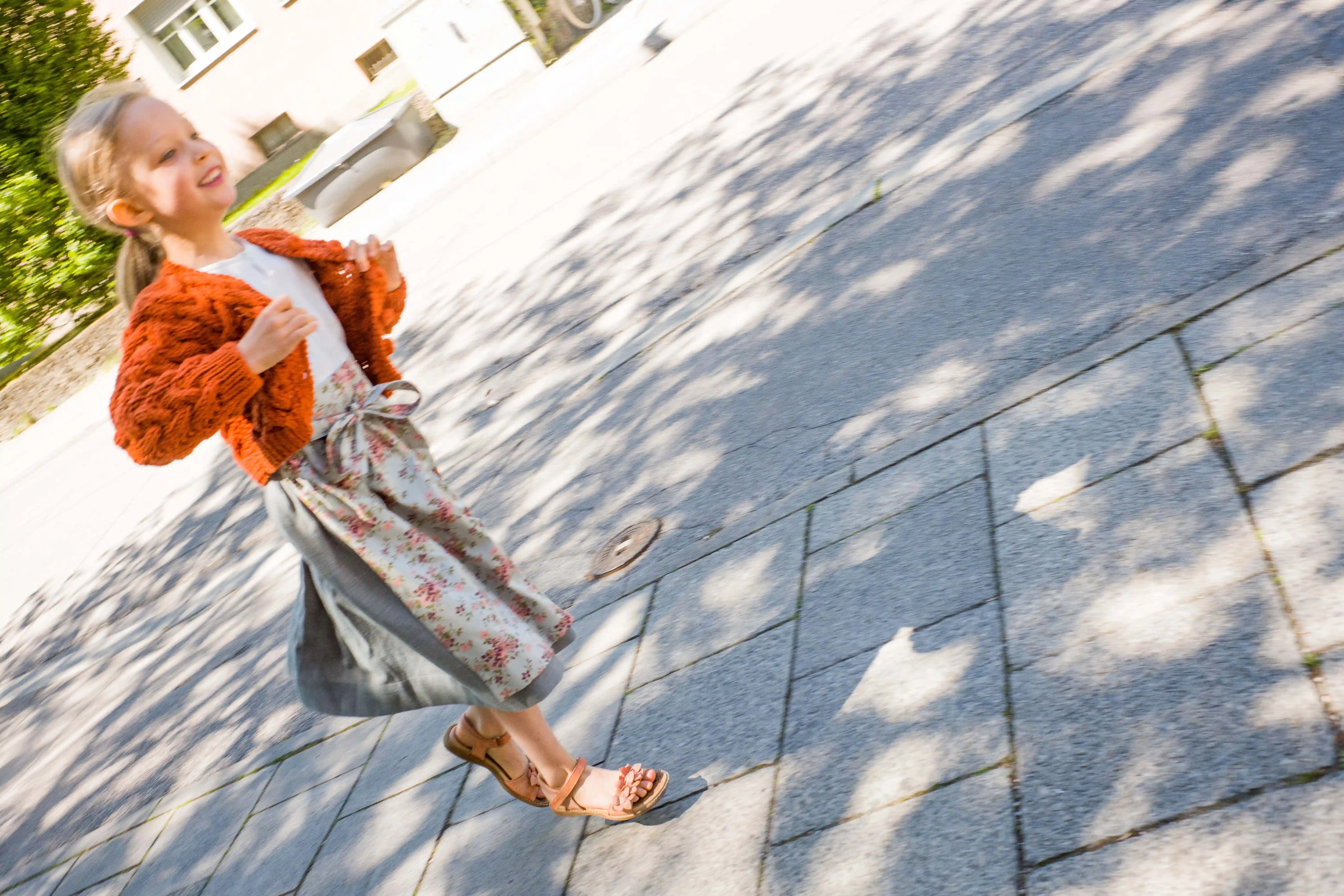 Zirbenfüxlein Kinderdirndl Saphir mit terrakottafarbener Zopf-Strickjacke und Blumenschürze seitliche Ansicht eines lachenden Kindes in Bewegung auf dem Gehweg mit Schattenspiel im urbanen Straßensetting, lebhaft interpretiert von Zirbenfüxlein.