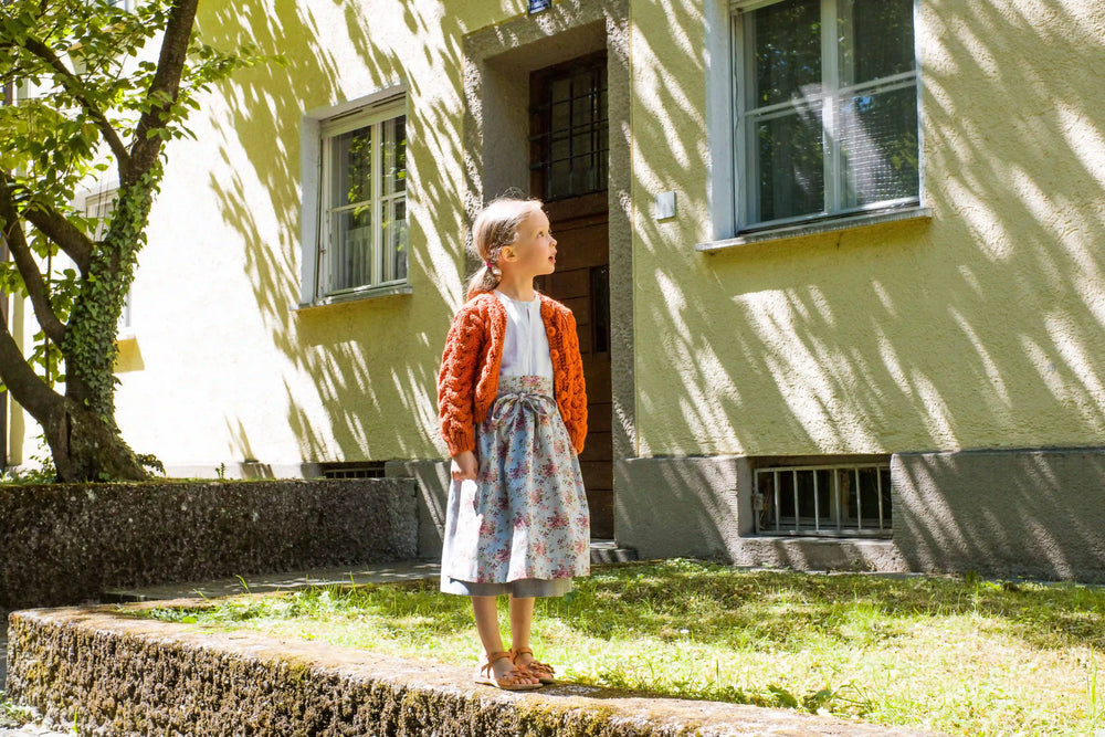 Zirbenfüxlein Kinderdirndl Saphir mit terrakottafarbener Zopf-Strickjacke, Frontansicht eines Kindes auf einem Mäuerchen stehend vor Häuserfassade mit sonnigem Lichtspiel in urbaner Szene, modern umgesetzt von Zirbenfüxlein.