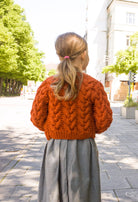 Zirbenfüxlein Kinderdirndl Saphir mit terrakottafarbener Zopf-Strickjacke, Nahaufnahme der Strickjacke in Rückansicht vor urbaner Straßenszene, hochwertig umgesetzt von Zirbenfüxlein.