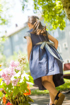 Zirbenfüxlein Kinderdirndl in Tintenblau, Rückansicht eines Kindes im Hintergrund in grüner Hinterhofszene, Blumen im scharfen Vordergrund hervorgehoben, ruhig interpretiert von Zirbenfüxlein.