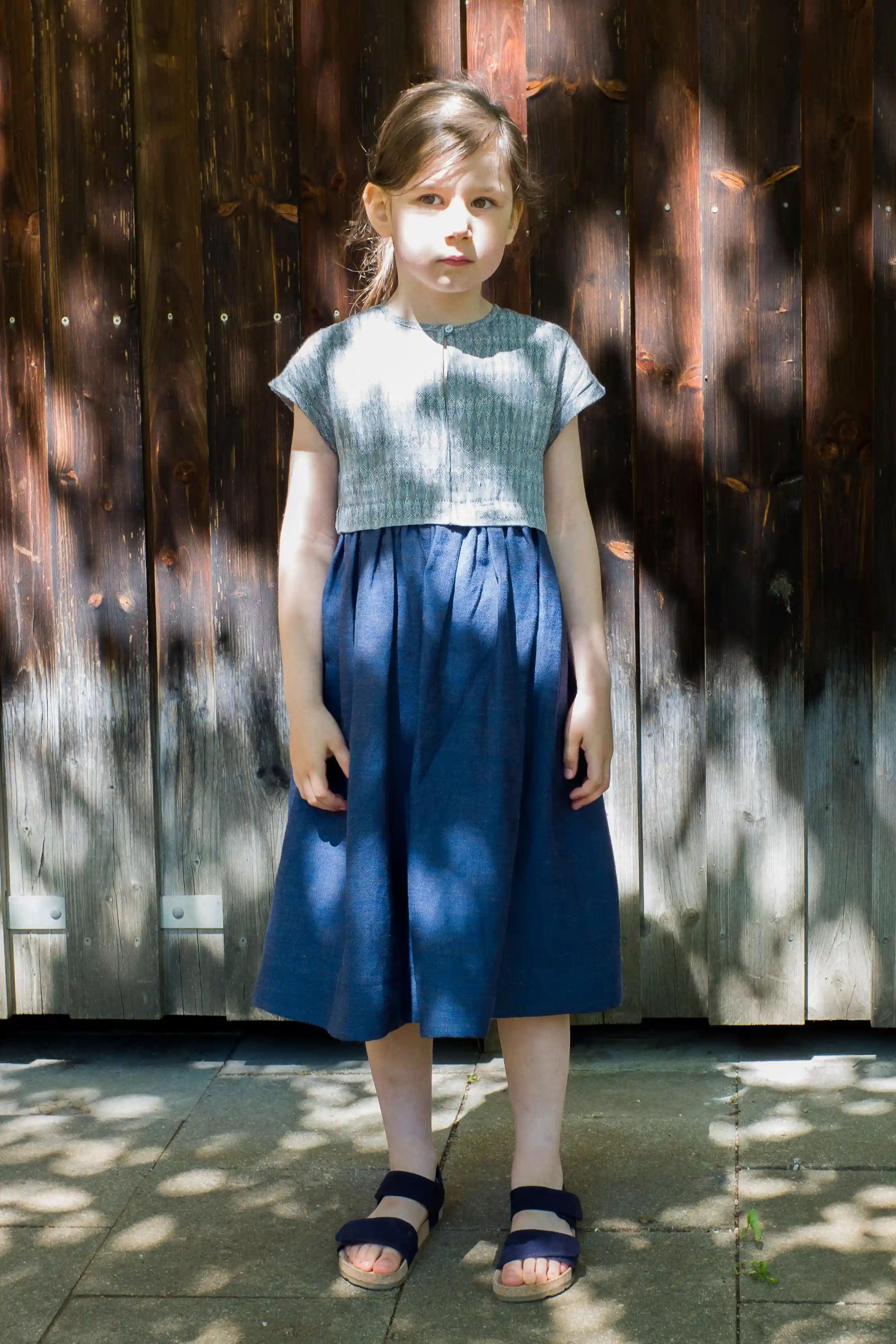 Kinderdirndl in Tintenblau aus Leinen-Schurwolle-Mischung – Frontansicht vor brauner Lattenwand mit Licht- und Schattenspiel, stimmungsvoll in Szene gesetzt von Zirbenfüxlein
