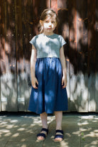 Kinderdirndl in Tintenblau aus Leinen-Schurwolle-Mischung – Frontansicht vor brauner Lattenwand mit Licht- und Schattenspiel, stimmungsvoll in Szene gesetzt von Zirbenfüxlein