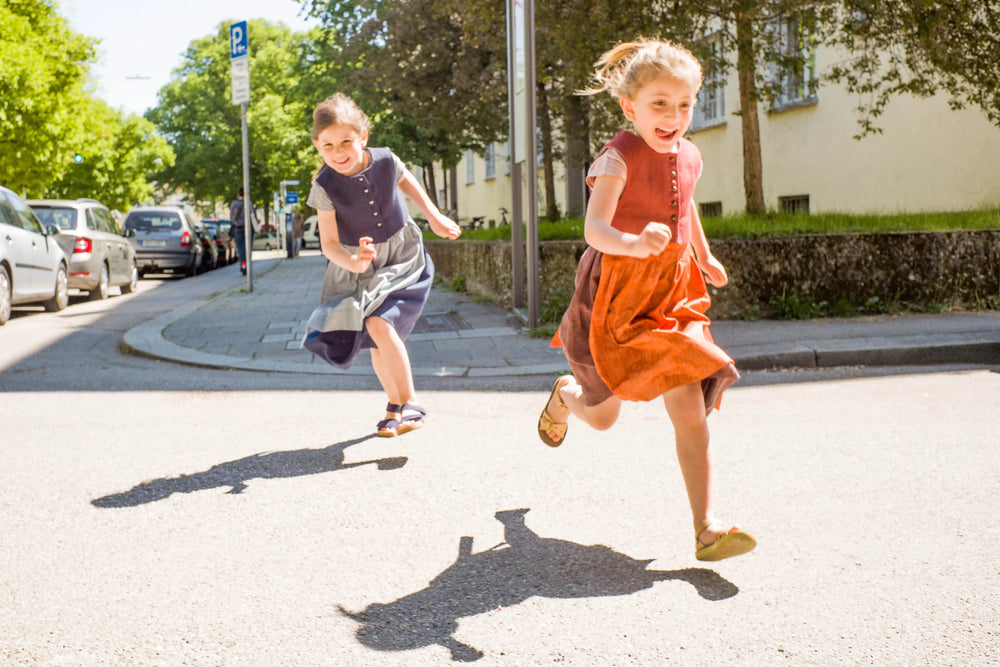 Zirbenfüxlein Kinderdirndl in Tintenblau und pinke Beere, zwei Kinder rennen lachend über eine sonnige Straße in urbaner Szene, dynamisch umgesetzt von Zirbenfüxlein.