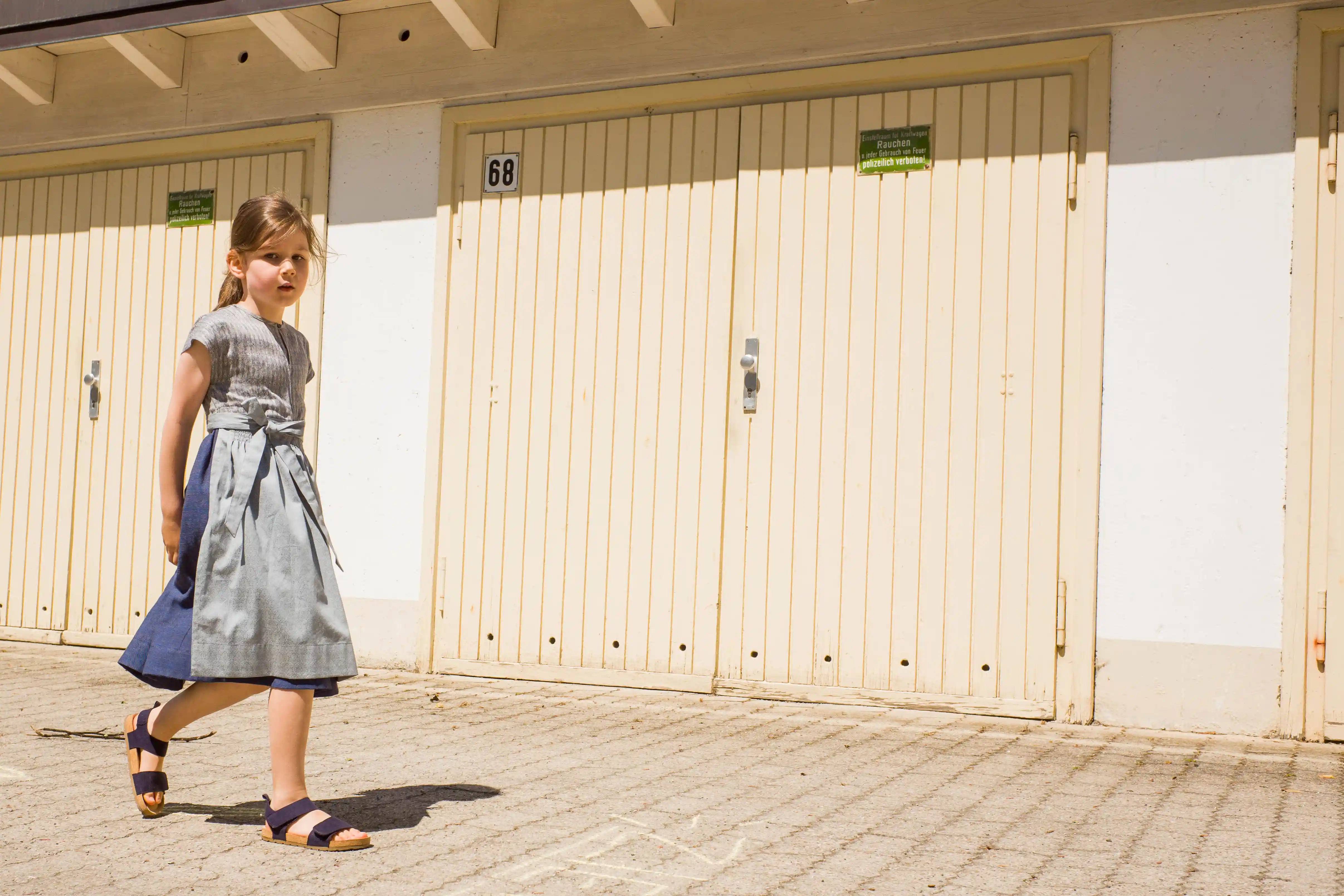 Leinen-Kinderdirndl in Tintenblau mit hellblauer Schürze – Einstellung vor Garagentor im Sonnenlicht, Sommermoment von Zirbenfüxlein