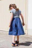 Leinen-Kinderdirndl in Tintenblau mit hellblauer Baumwollchürze – Rückansicht beim Drehen vor Garageneinfahrt, verspielte Bewegung, Kind blickt nach unten, Szene von Zirbenfüxlein