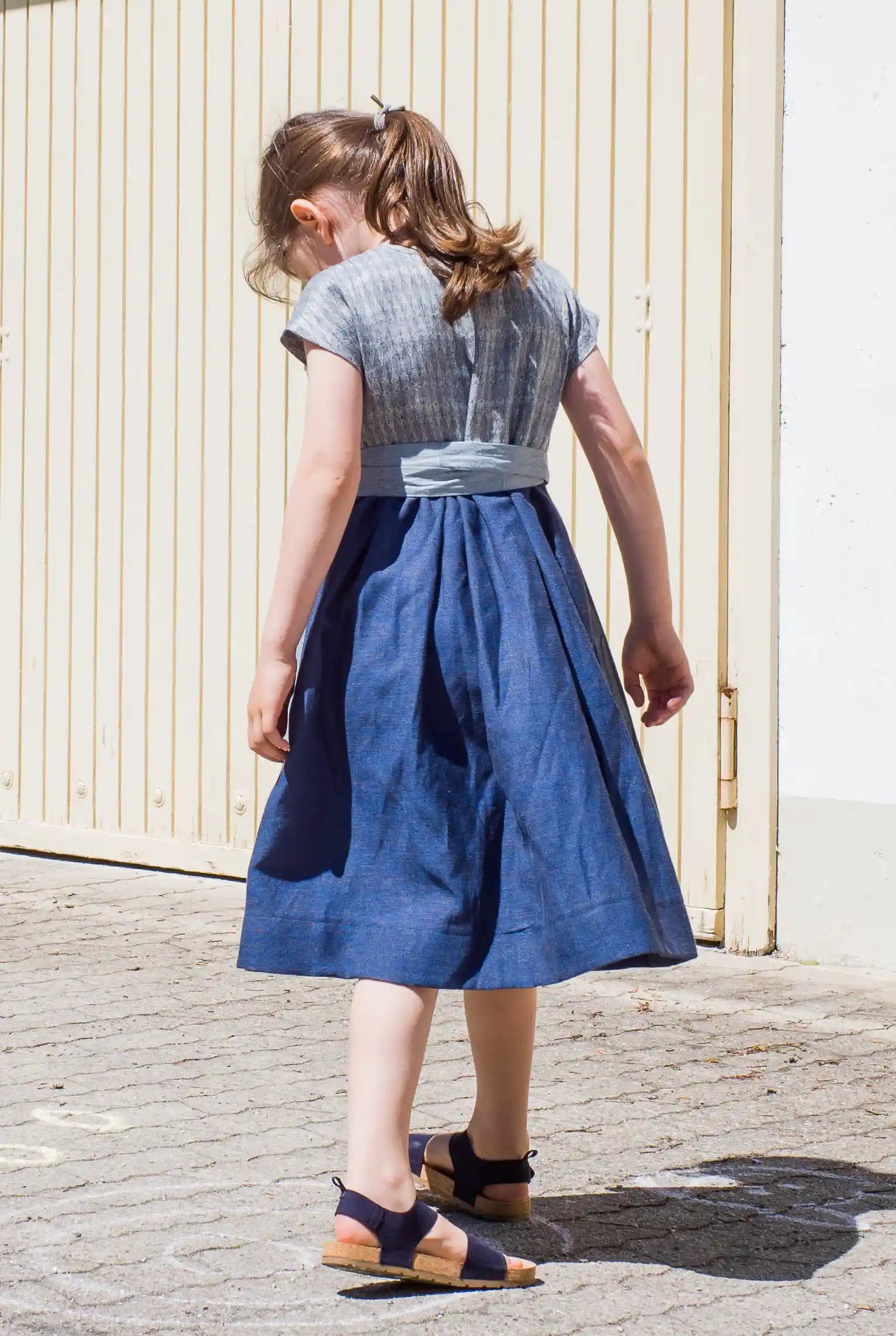 Leinen-Kinderdirndl in Tintenblau mit hellblauer Baumwollchürze – Rückansicht beim Drehen vor Garageneinfahrt, verspielte Bewegung, Kind blickt nach unten, Szene von Zirbenfüxlein