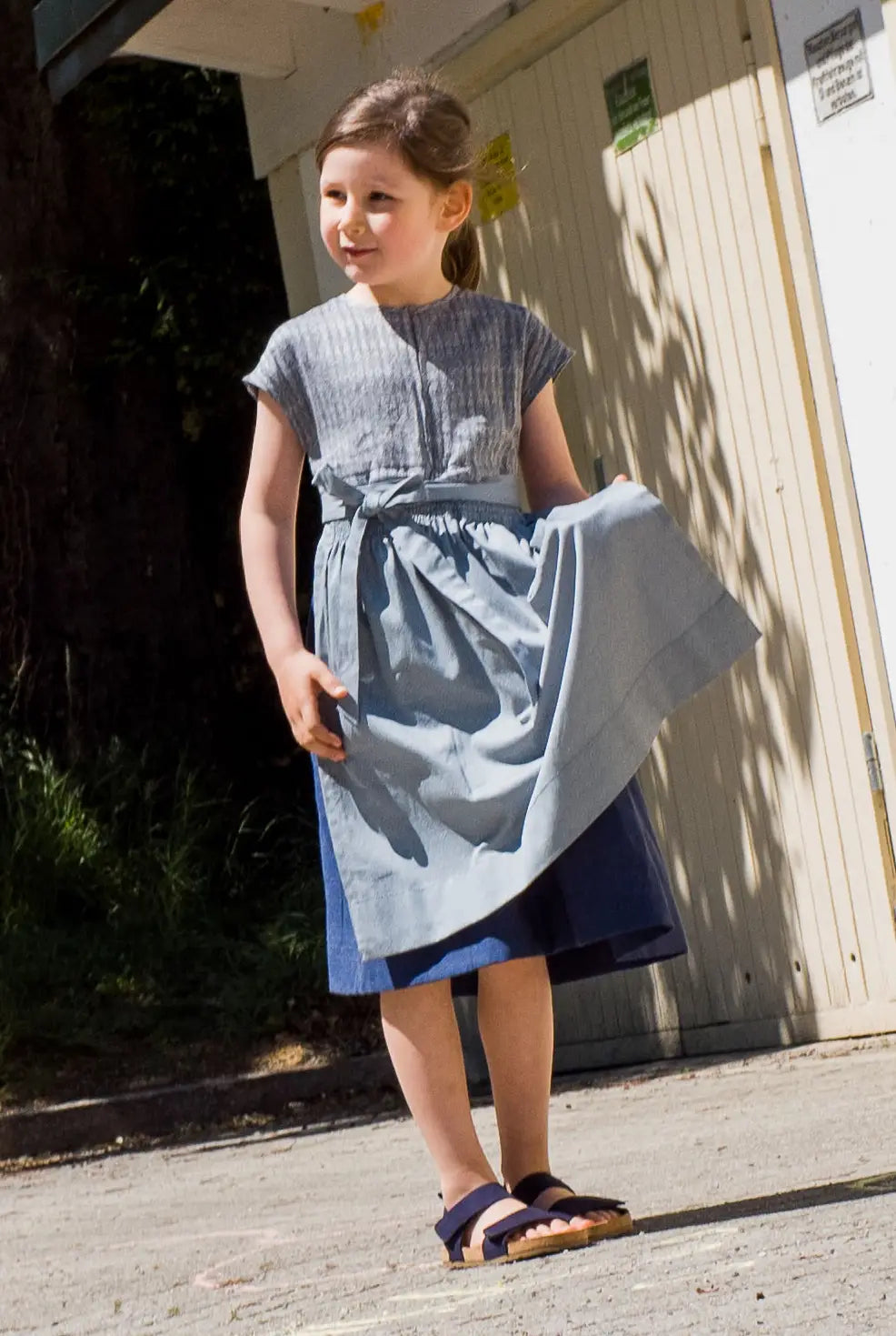 Leinen-Kinderdirndl in Tintenblau mit hellblauer Baumwollschürze – stehend vor Garagentor, das Kind hält die Schürze in der Hand, inszeniert von Zirbenfüxlein