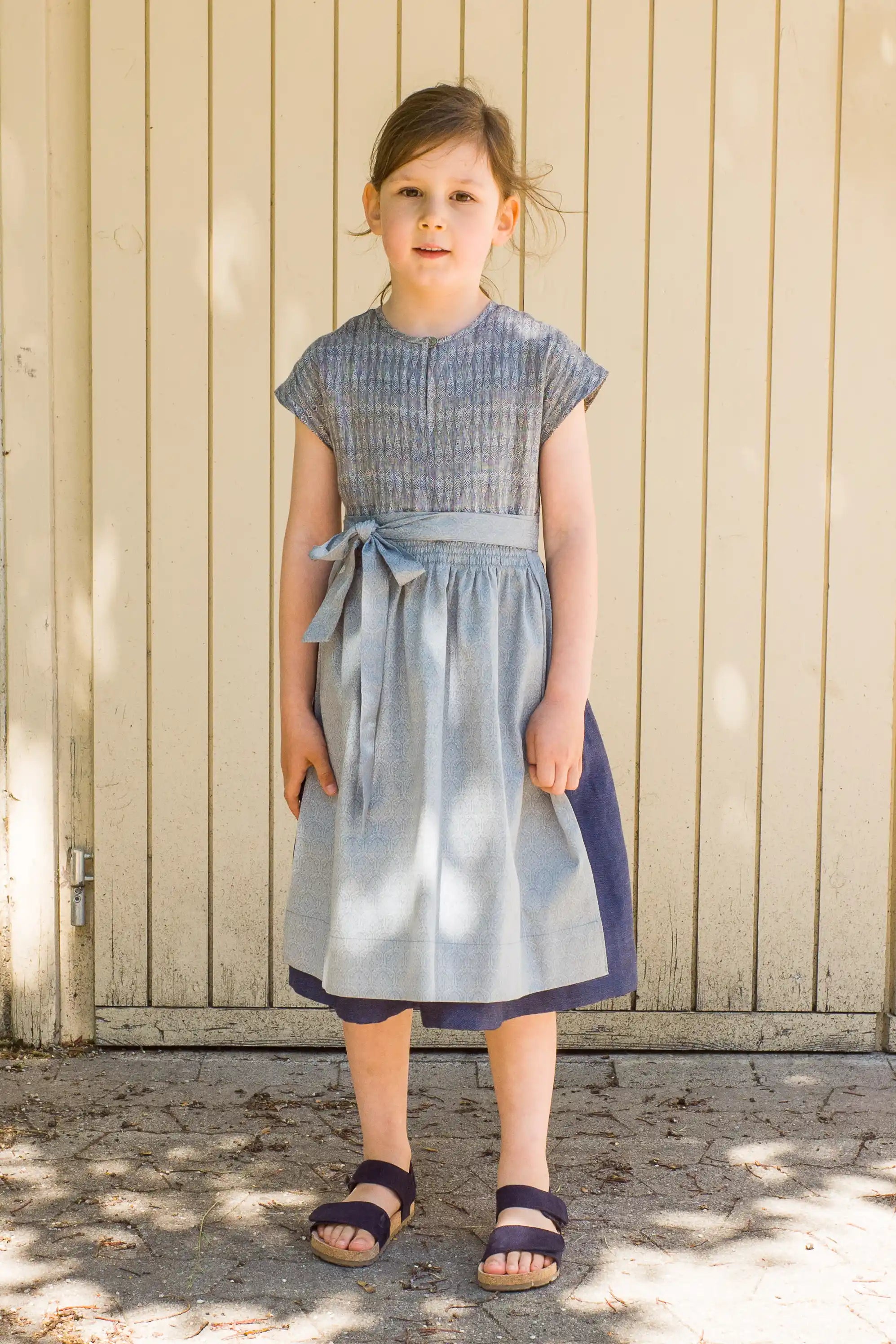 Leinen-Kinderdirndl in Tintenblau mit hellblauer Baumwollschürze – Frontansicht vor Garagentor, modern inszeniert von Zirbenfüxlein