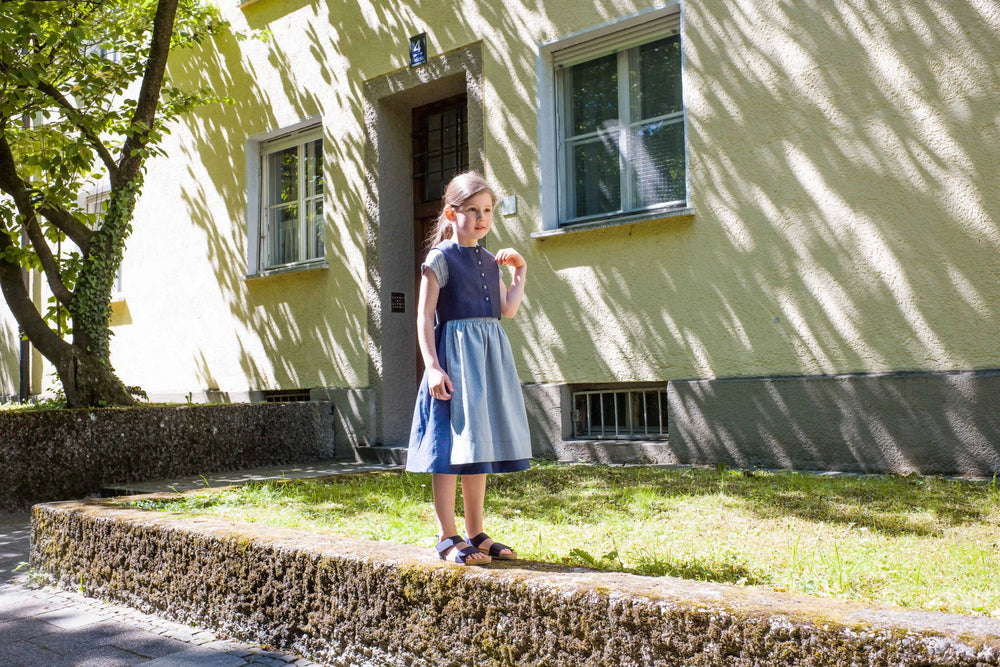 Kinderdirndl in Tintenblau mit hellblauer Schürze – Kind steht seitlich auf einem Mäuerchen, im Hintergrund urbane Häuserfassade mit Licht- und Schattenspiel, inszeniert von Zirbenfüxlein