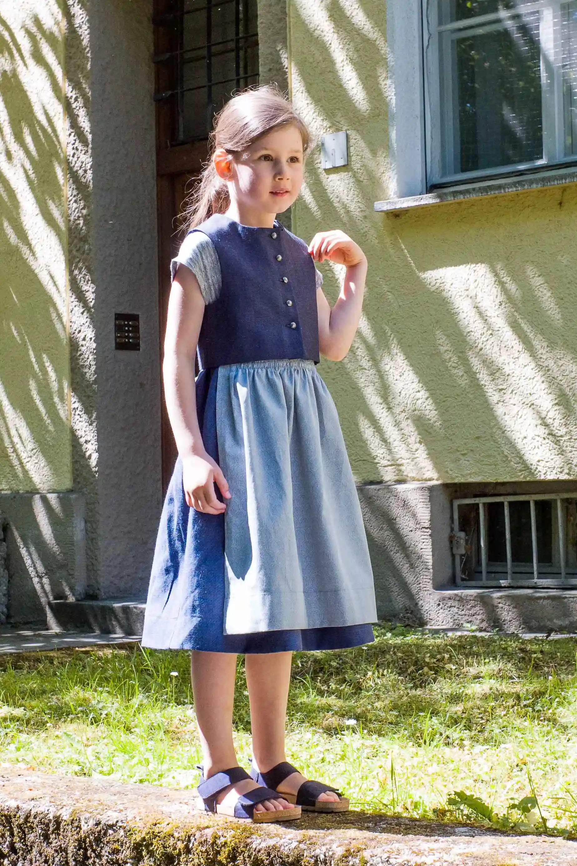Kinderdirndl in Tintenblau mit hellblauer Schürze – Kind steht seitlich auf einem Mäuerchen, im Hintergrund urbane Häuserfassade mit Licht- und Schattenspiel, freundlich inszeniert von Zirbenfüxlein