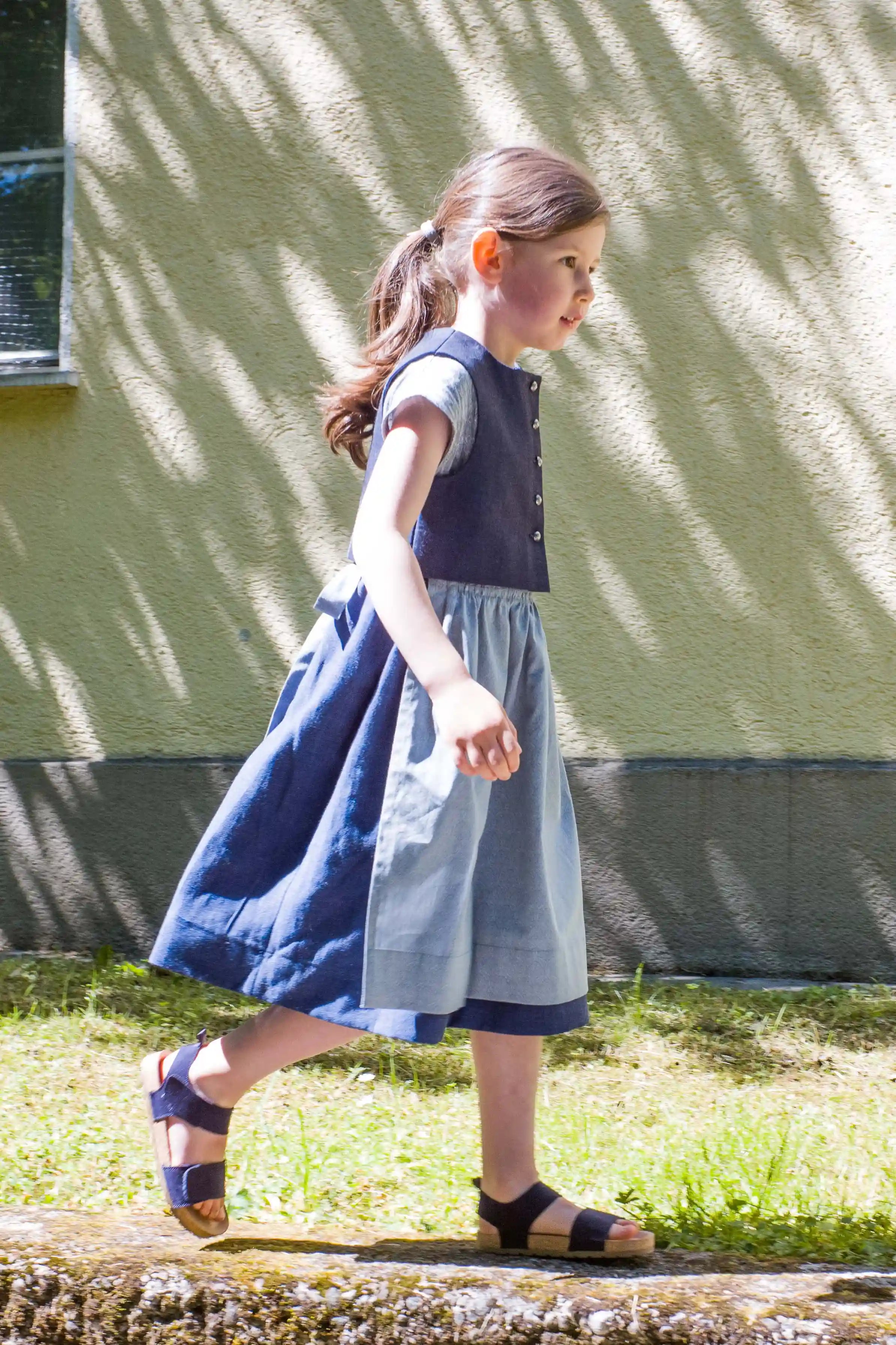 Kinderdirndl in Tintenblau mit hellblauer Schürze – Kind läuft von links nach rechts auf einem Mäuerchen entlang, vor urbaner Häuserfassade mit Licht- und Schattenspiel, lebendig inszeniert von Zirbenfüxlein