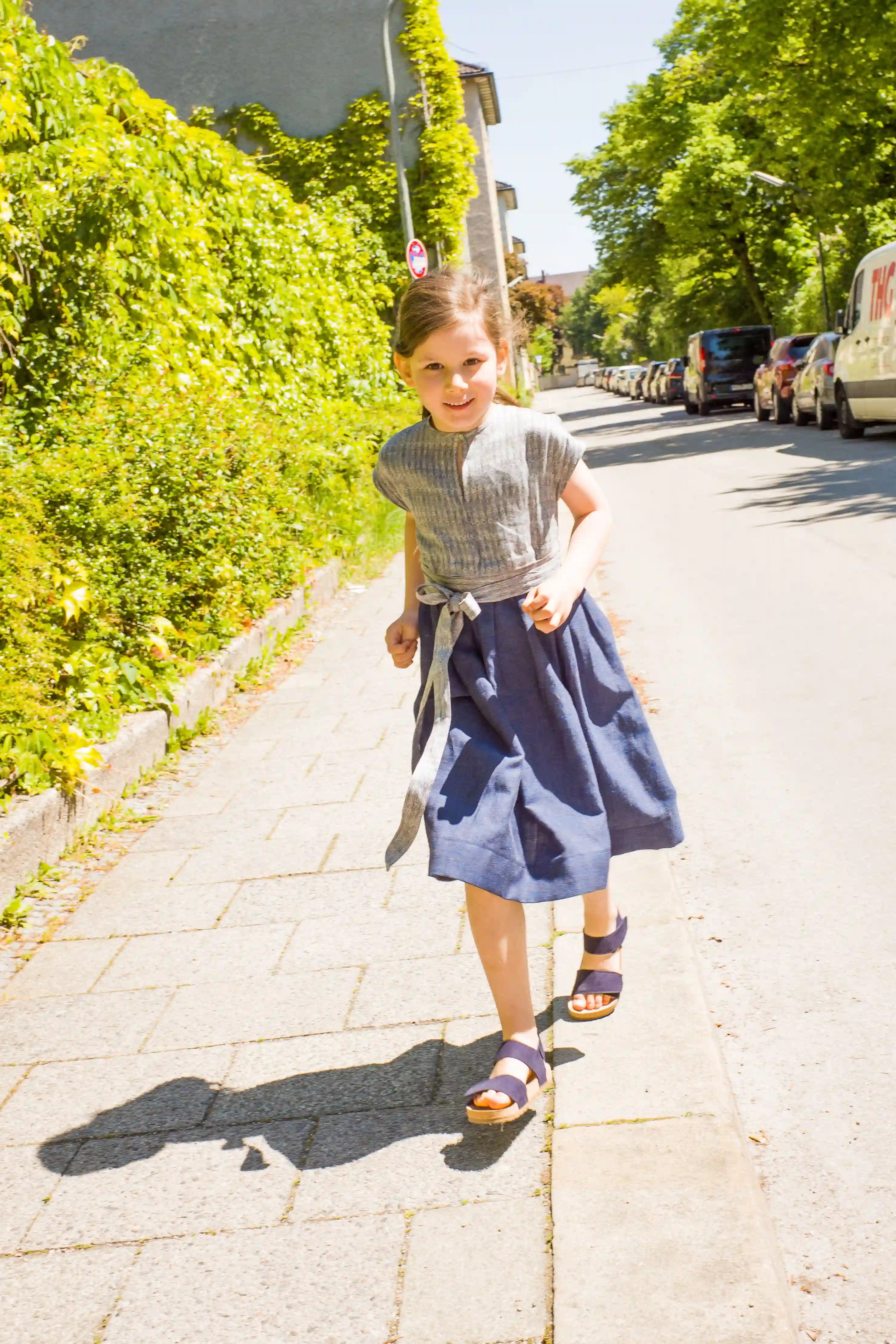 Kinderdirndl in Tintenblau mit Schürzenband – Kind läuft auf Gehweg entlang einer Straße im urbanen Setting, lebendig inszeniert von Zirbenfüxlein