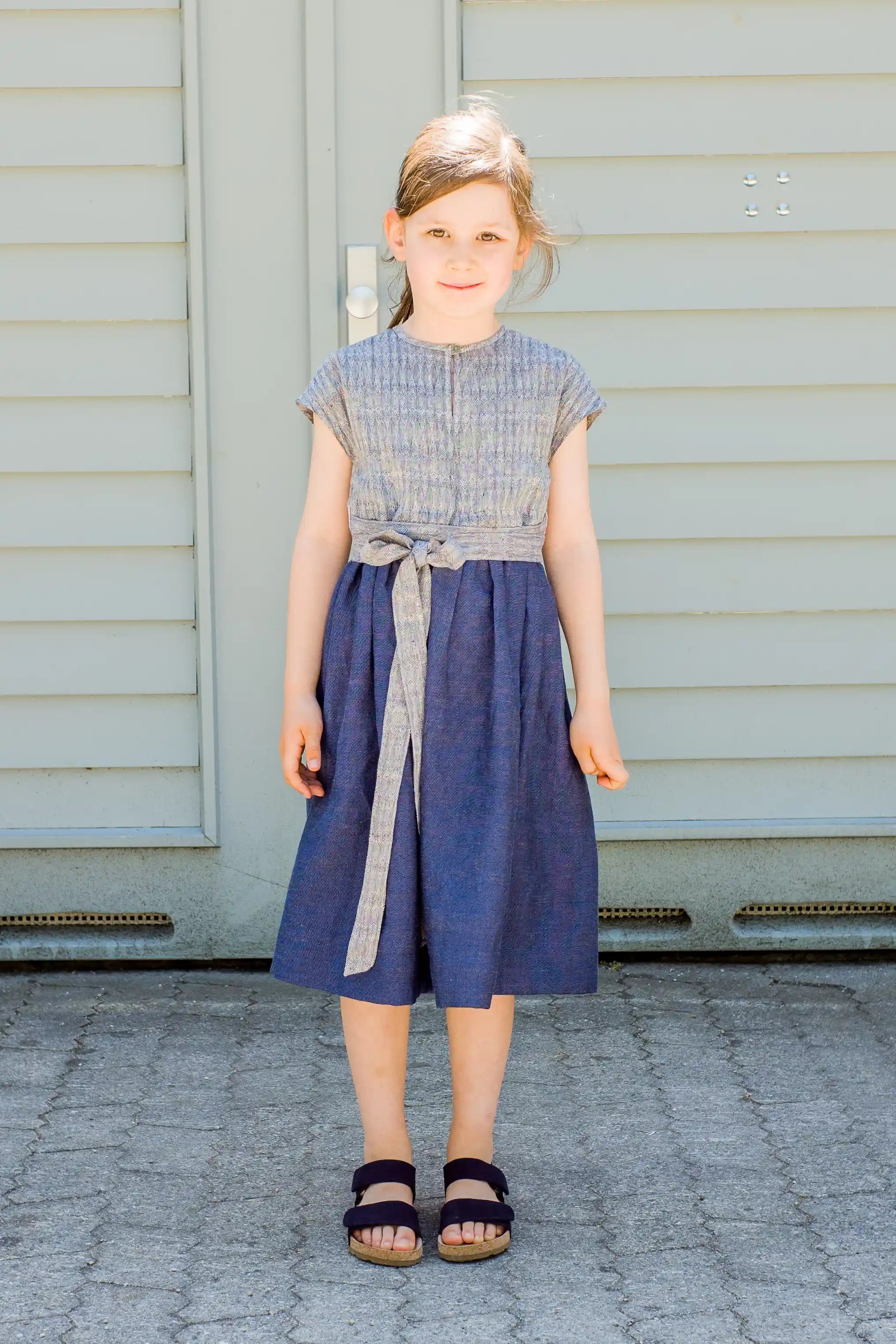 Kinderdirndl in Tintenblau mit Schürzenband – Kind steht frontal vor einem Garagentor, klar und reduziert inszeniert von Zirbenfüxlein