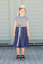 Kinderdirndl in Tintenblau mit Schürzenband – Kind steht frontal vor einem Garagentor, klar und reduziert inszeniert von Zirbenfüxlein