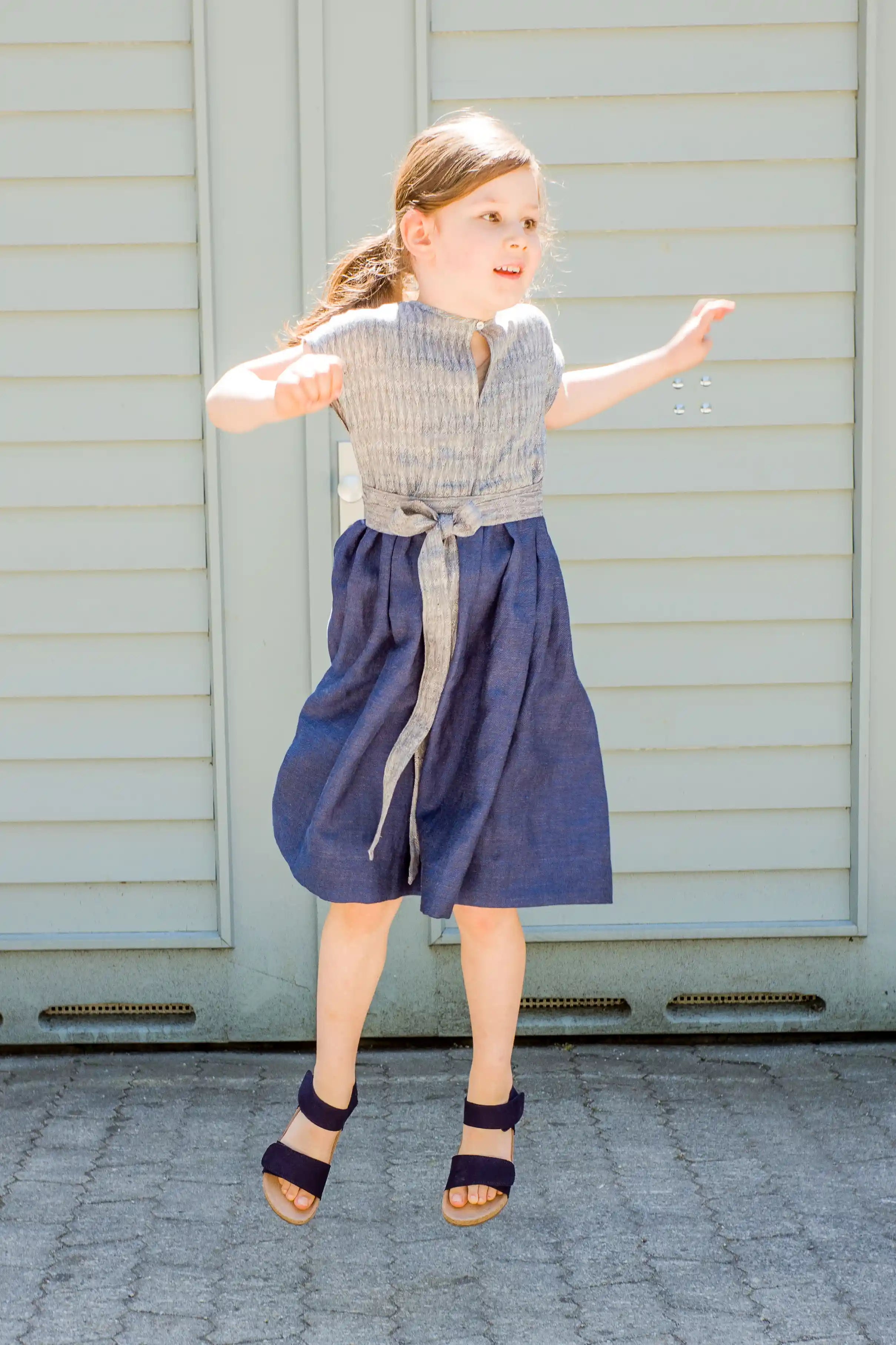 Kinderdirndl in Tintenblau mit Schürzenband – Kind springt vor einem Garagentor nach oben, fröhlich inszeniert von Zirbenfüxlein