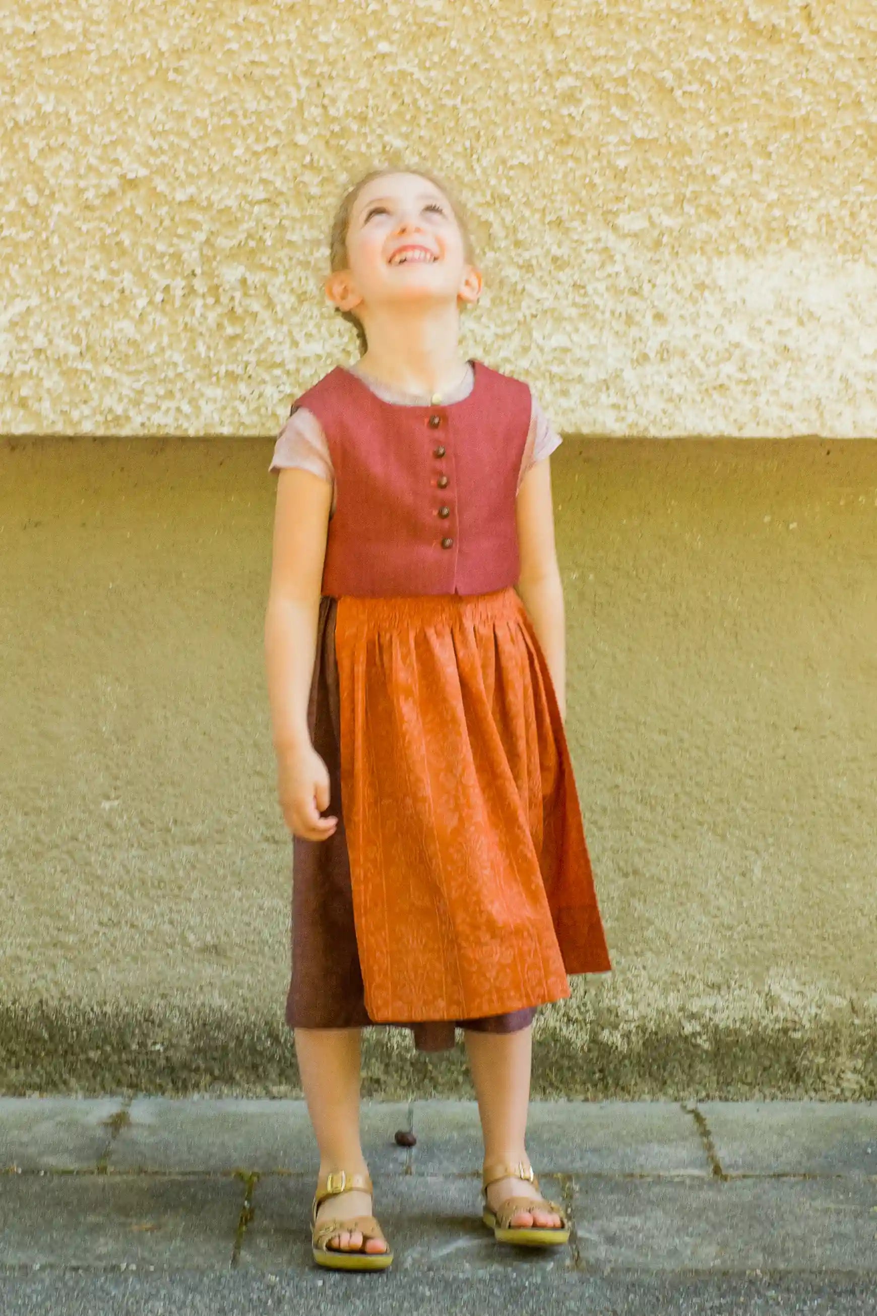 Kinderdirndl in pinker Beere mit terrakottafarbener Schürze und Weste – stehend vor urbaner Fassade mit Blick nach oben, inszeniert von Zirbenfüxlein