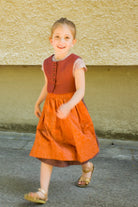 Kinderdirndl in pinker Beere mit terrakottafarbener Schürze und Weste – nach vorne aus dem Bild laufend vor urbaner Fassade, lebendig und modern inszeniert von Zirbenfüxlein