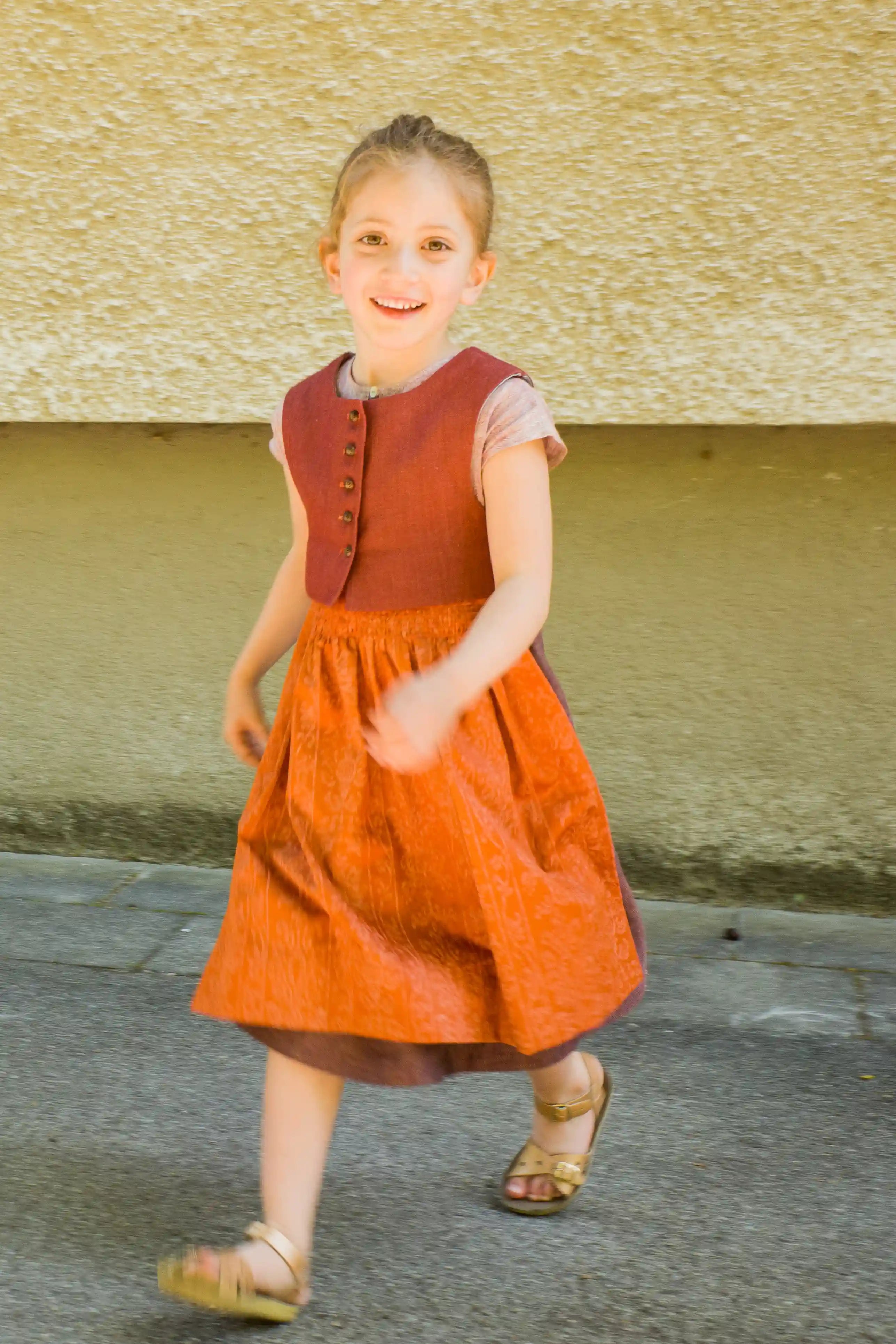Kinderdirndl in pinker Beere mit terrakottafarbener Schürze und Weste – nach vorne aus dem Bild laufend vor urbaner Fassade, lebendig und modern inszeniert von Zirbenfüxlein