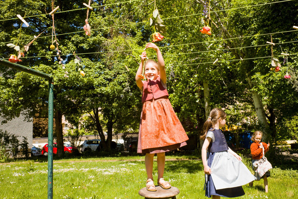 Kinderdirndl in Pink mit terrakottafarbener Schürze und Weste – stehend auf einem alten Hocker auf einer Hinterhofwiese, Blumen von einer Wäscheleine abnehmend, zwei weitere Kinder im Hintergrund, spielerisch inszeniert von Zirbenfüxlein
