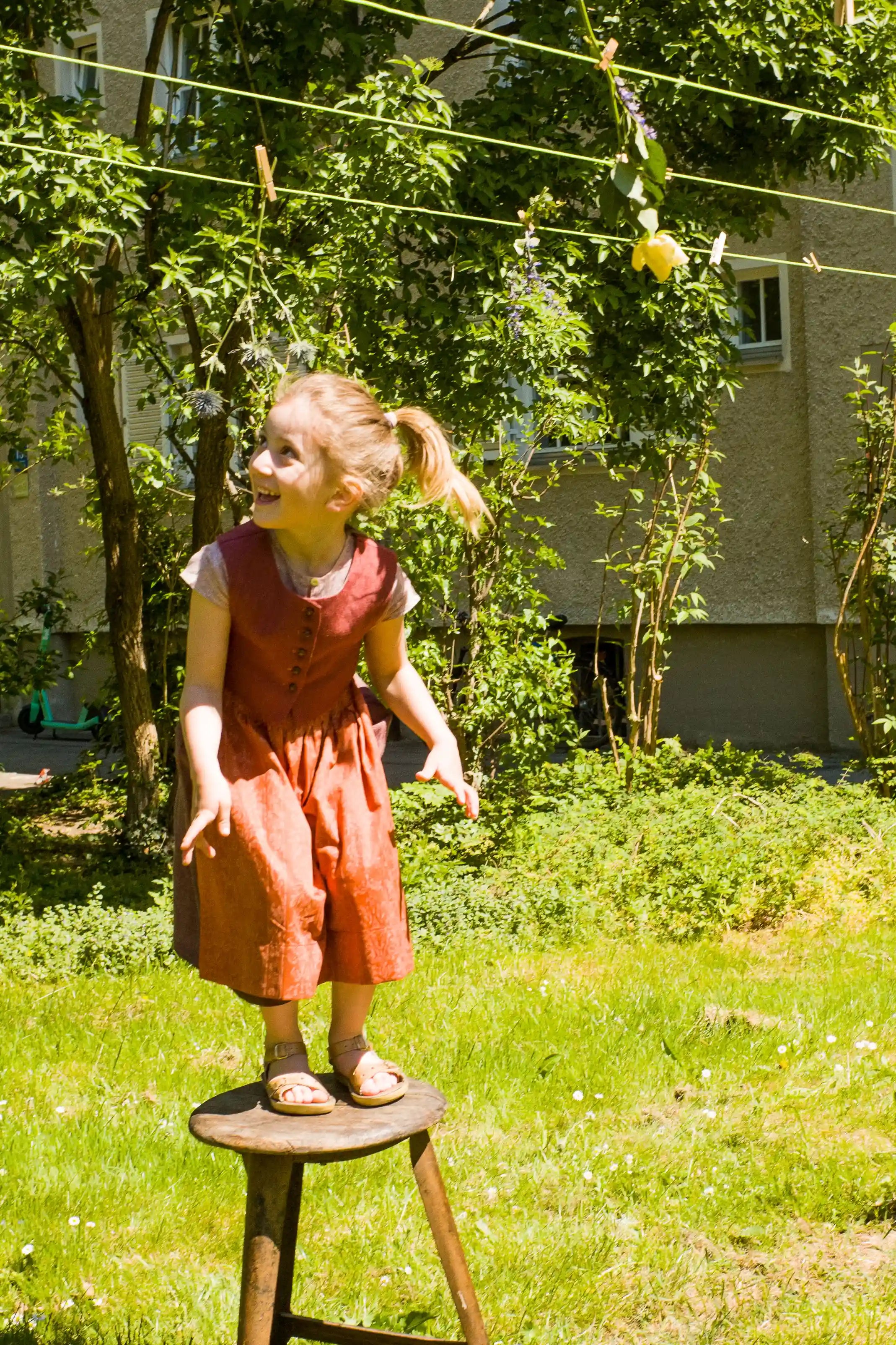 Kinderdirndl in Pink mit terrakottafarbener Schürze und Weste – in einer Wiese in einem Hinterhof auf einem alten Hocker stehend, Blick nach oben zu einer Wäscheleine, inszeniert von Zirbenfüxlein