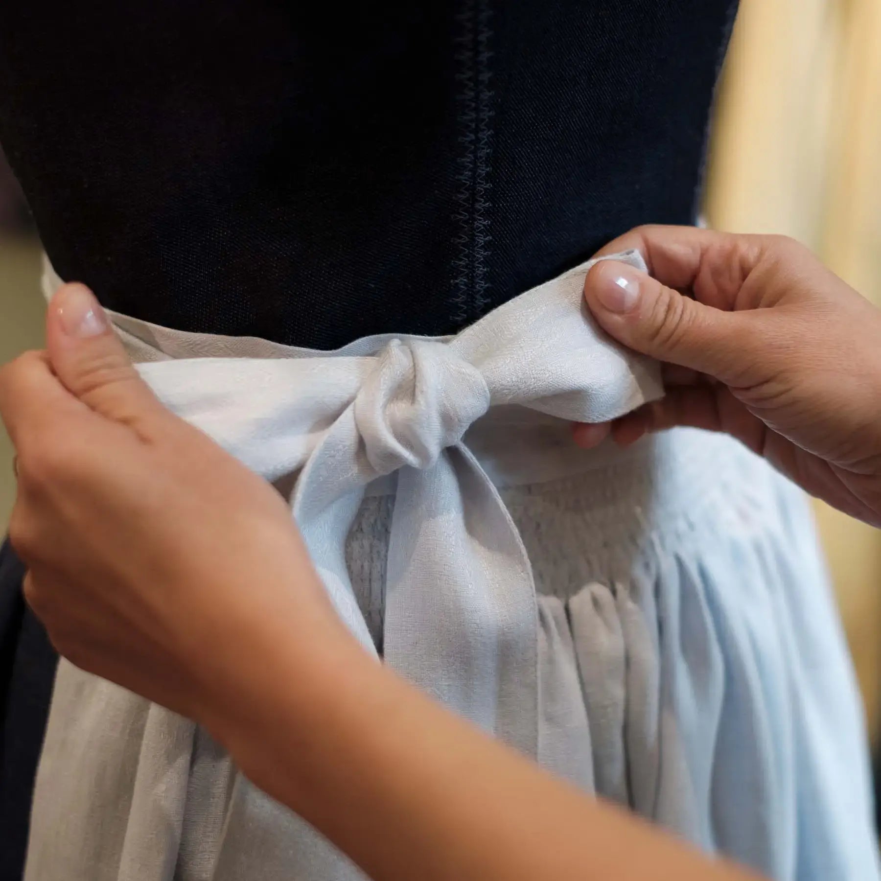 Zirbenfux Atelier München, Nahaufnahme einer Dirndlschleife, die gerade fertig gebunden wird, konzentriert und handwerklich dargestellt.