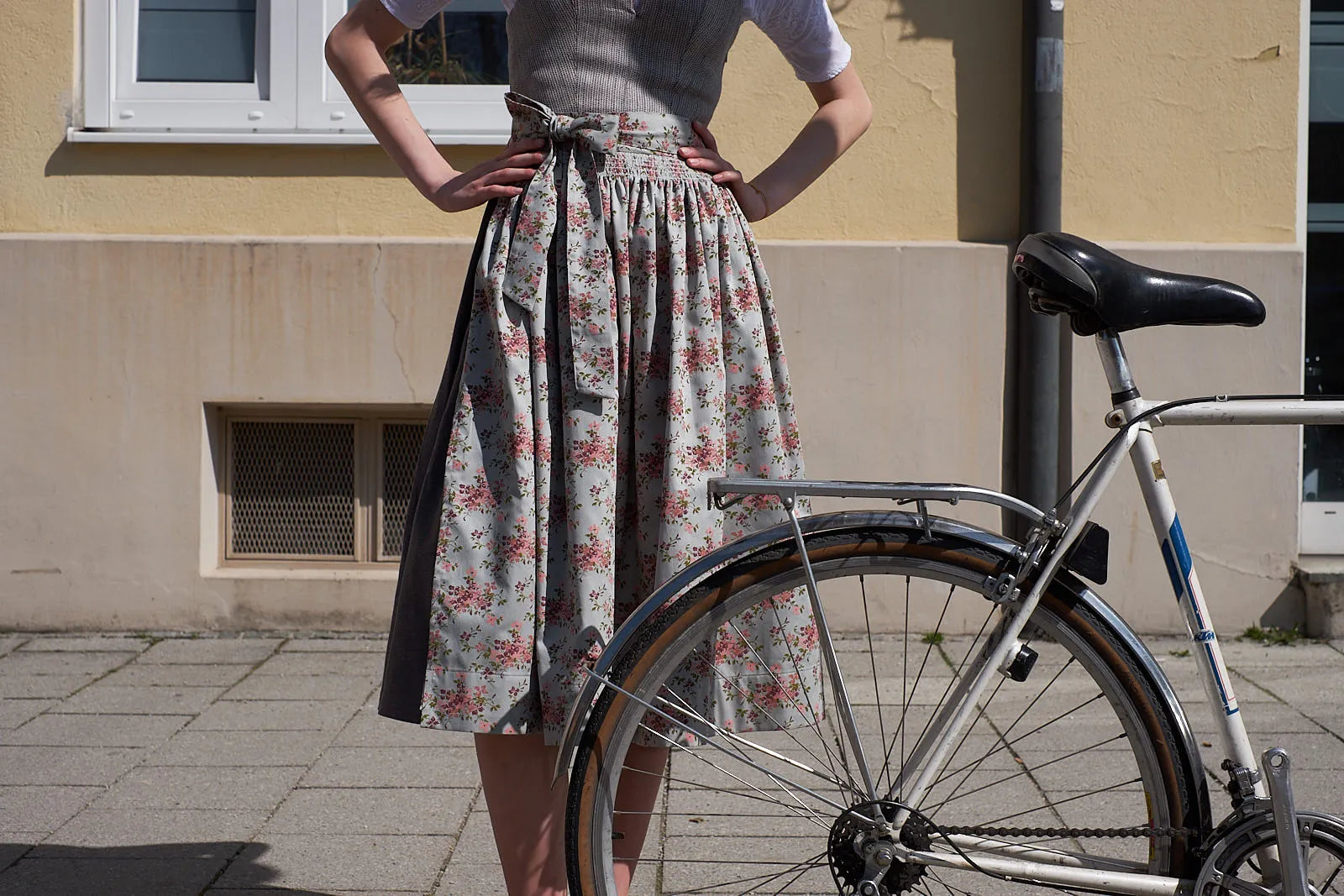 Dirndlschürze mit floralem Muster im Zentrum – Model im Anschnitt auf Gehweg, neben einem alten Rennrad, urban in Szene gesetzt von Zirbenfux