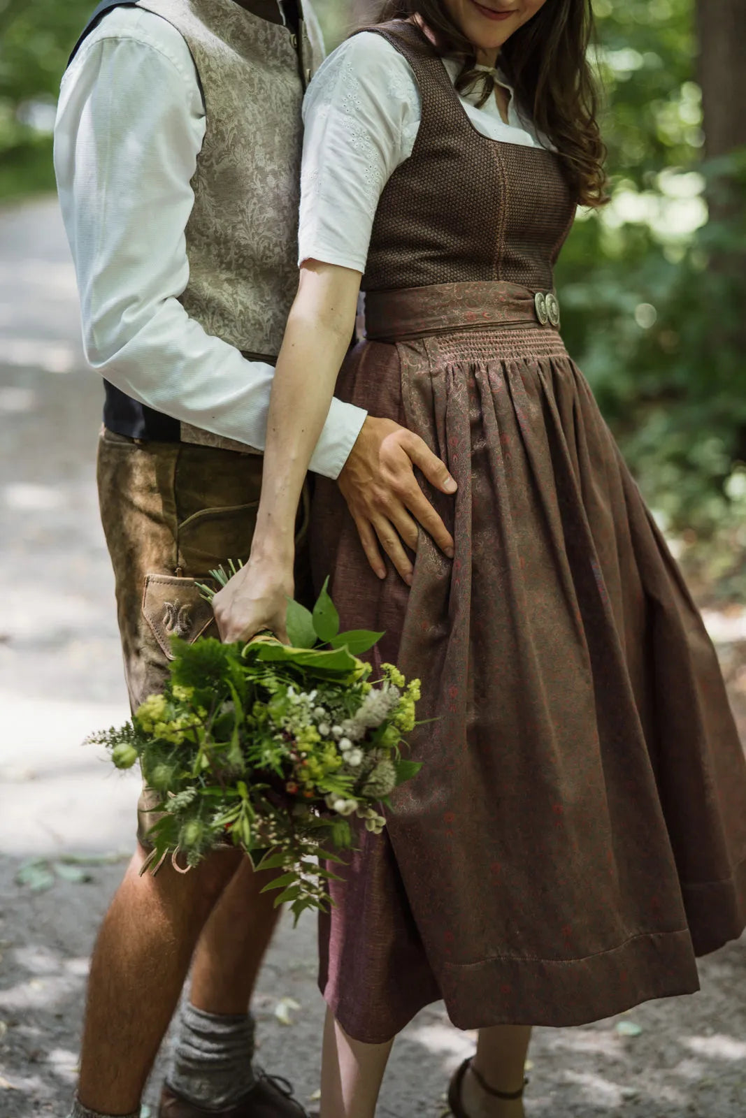 Zirbenfux Hochzeitsdirndl der Braut in Beerentönen, Brautpaar im Park vertraut hintereinander stehend dargestellt, ruhig komponiert von Zirbenfux.