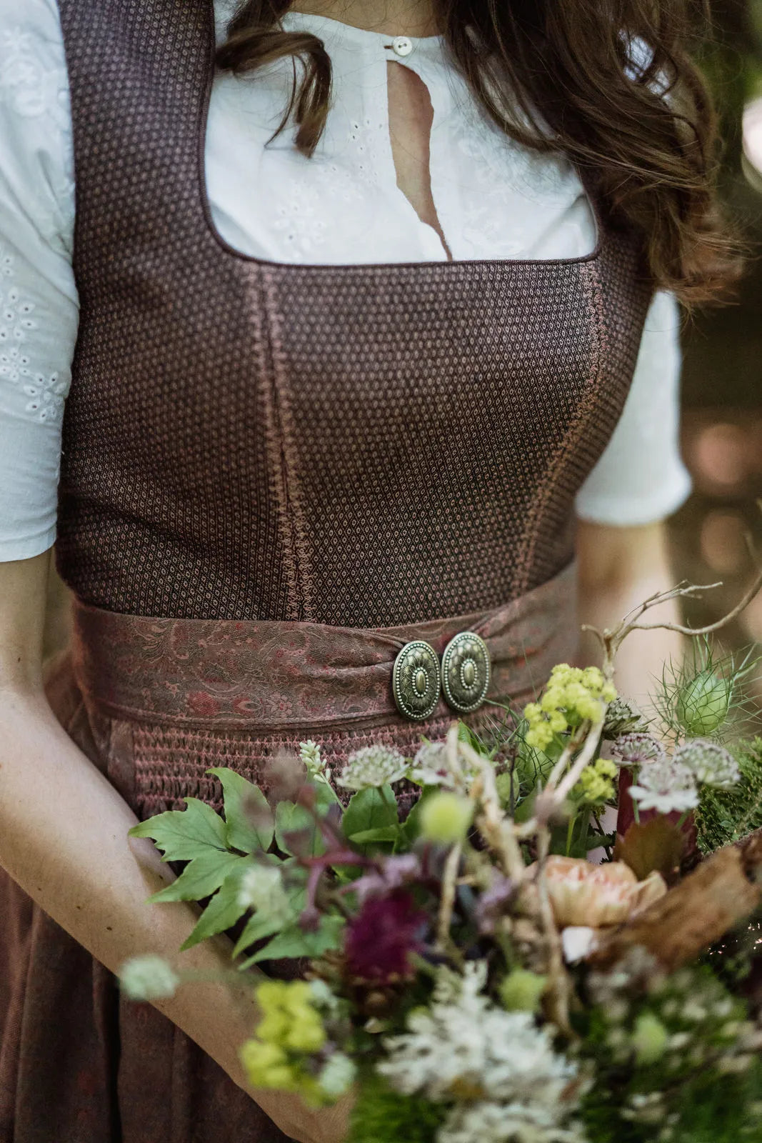 Zirbenfux Hochzeitsdirndl der Braut in Beerentönen, Frontansicht im Anschnitt mit gehaltenem Brautstrauß in urbaner Parks­zene, klar inszeniert von Zirbenfux.
