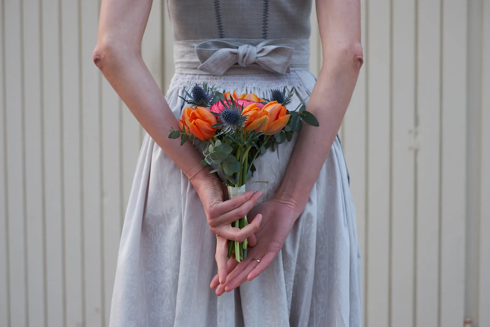 Zirbenfux Braut mit Hochzeitsdirndl in Silber, Ausschnitt in Rückansicht mit Brautstrauß auf Hüfthöhe hinter dem Rücken mit beiden Händen gehalten, Garagentor im Hintergrund sichtbar, komponiert inszeniert.