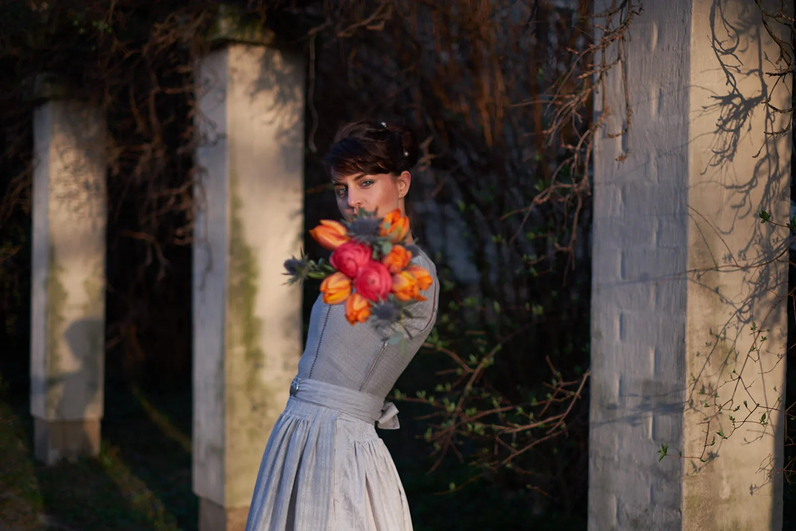 Zirbenfux mit Braut in Hochzeitsdirndl in Silber, Model streckt den Brautstrauß frontal in Richtung Kamera und blickt dahinter leicht provokant daran vorbei, selbstbewusst inszeniert.