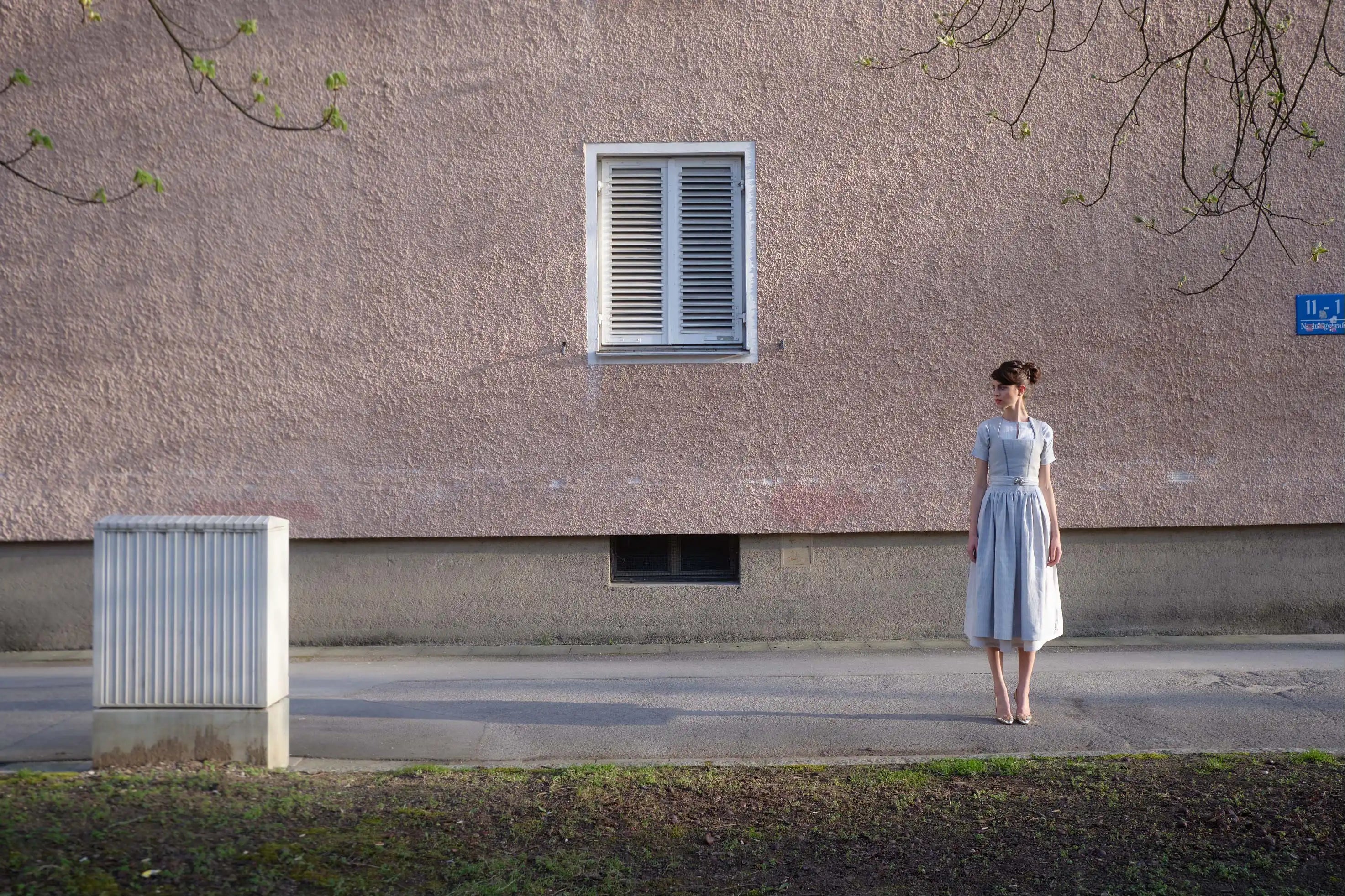 Zirbenfux Hochzeitsdirndl, Braut in Silber, Aufnahme aus der Weite eines Models ruhig und gerade vor urbaner Hausfassade im Abendlicht stehend mit Blick nach rechts, zurückgenommen inszeniert.