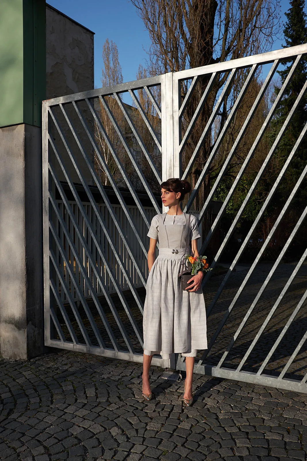 Zirbenfux Braut in Silber Hochzeitsdirndl, Frontansicht in Abendstimmung, Model an ein schmiedeeisernes Einfahrtstor angelehnt auf Kopfsteinpflaster stehend, Blick in die Ferne, Blumen locker in einer Tasche in der Hand, atmosphärisch inszeniert.