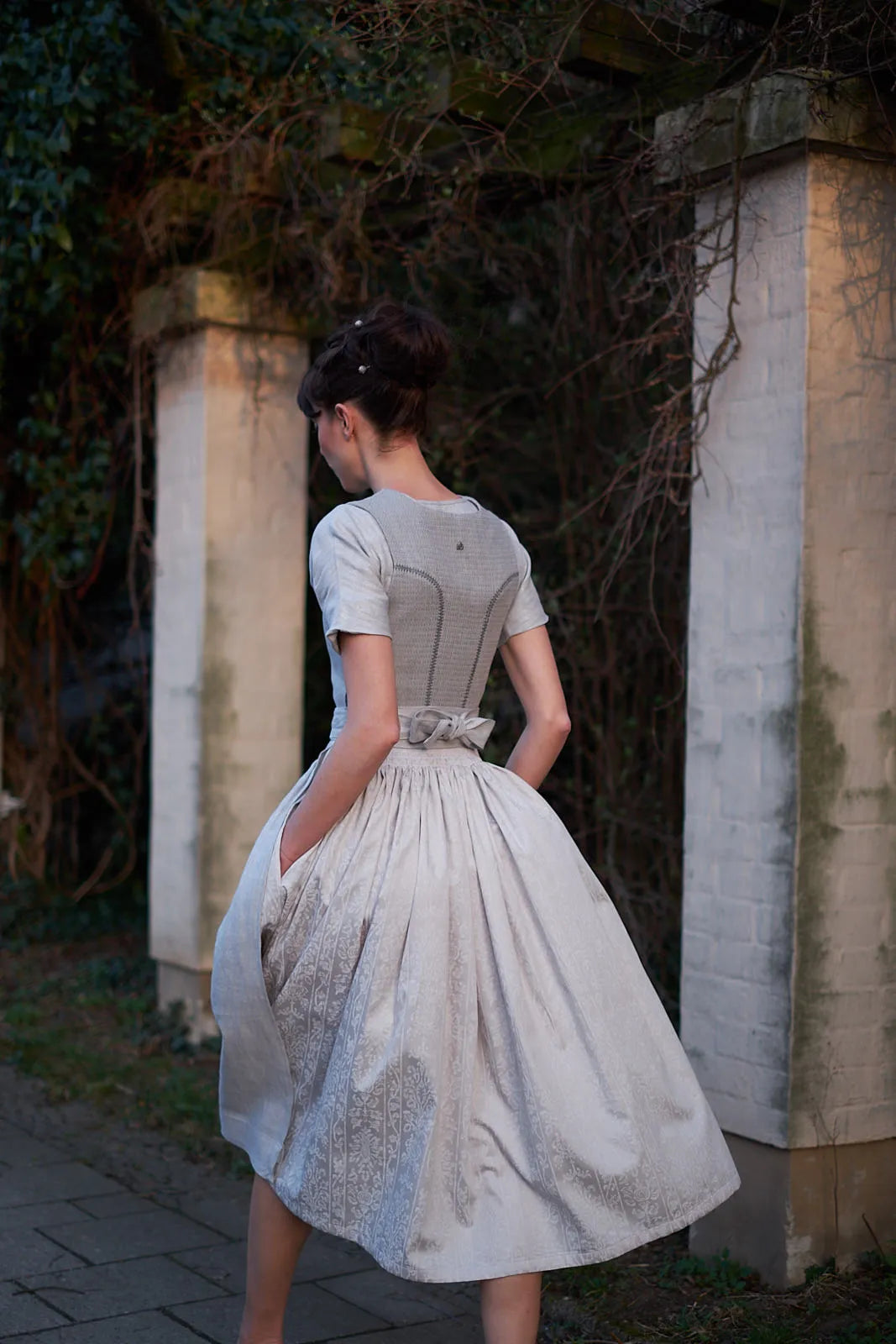 Zirbenfux Hochzeitsdirndl der Braut in Silber, Rückansicht eines Models eine Straße entlanggehend vom Betrachter weg, Rock schwingt weit und dynamisch im urbanen Umfeld, kraftvoll inszeniert von Zirbenfux.