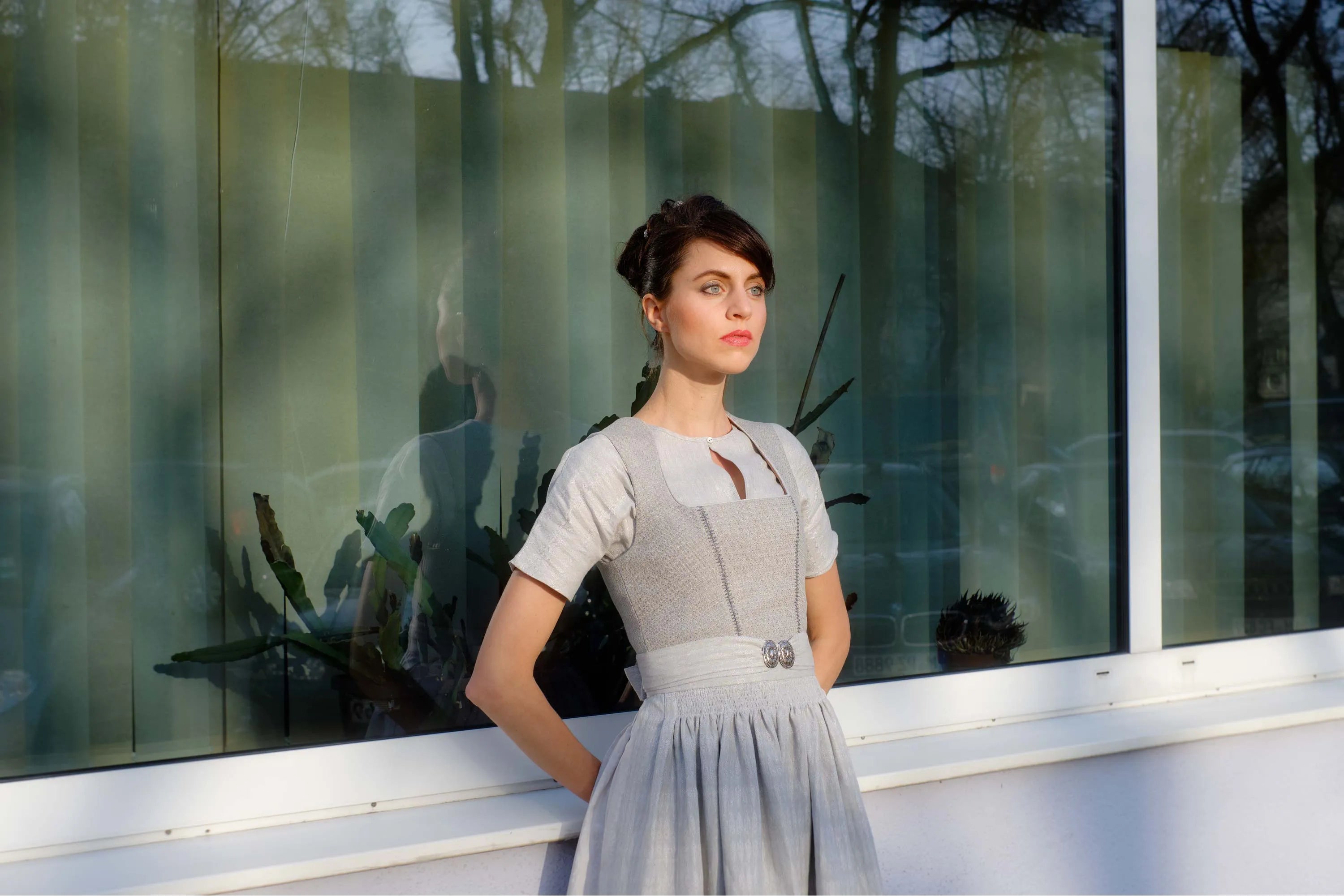 Zirbenfux Hochzeitsdirndl der Braut in Silber, Ansicht eines unbewegten Models mit dem Rücken an eine Fensterfront gelehnt in urbanem Setting, reduziert interpretiert von Zirbenfux.